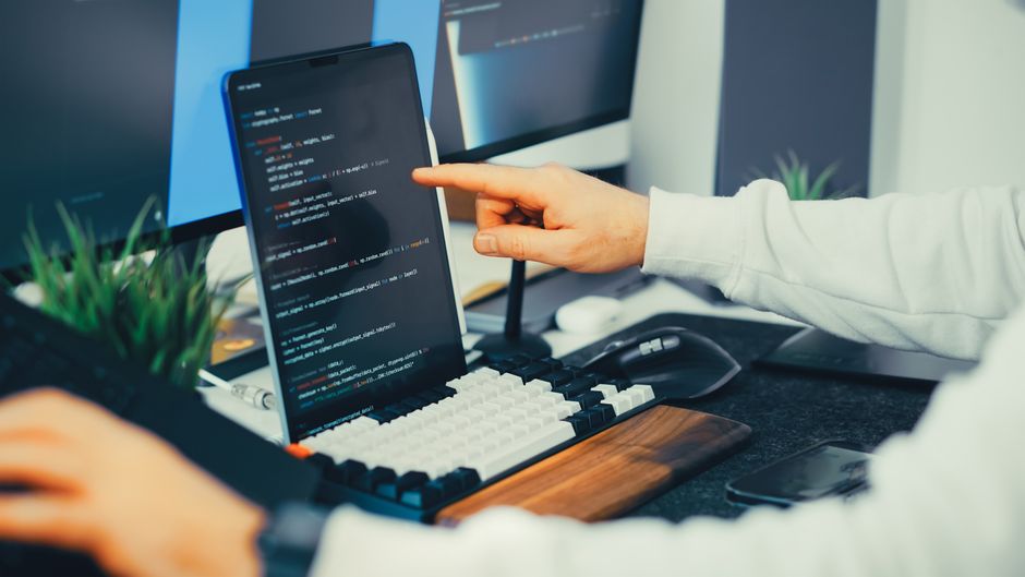 A person pointing at code on a tablet screen while another types on a keyboard