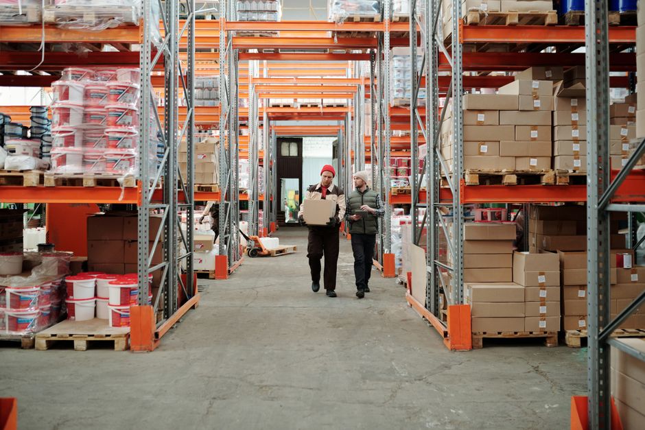 Two workers walking down a warehouse aisle between shelves of boxes, one carrying a parcel