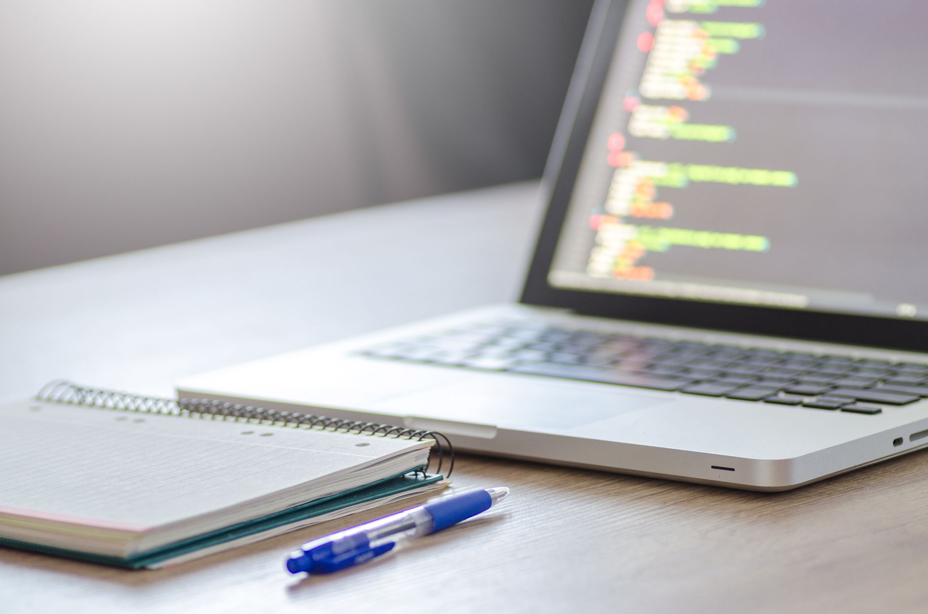 An open laptop showing code on screen, with a notebook and a blue pen on the desk