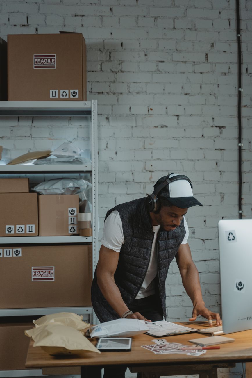 A man in headphones checking paperwork at a desk beside a computer, with shelves of boxed parcels behind him
