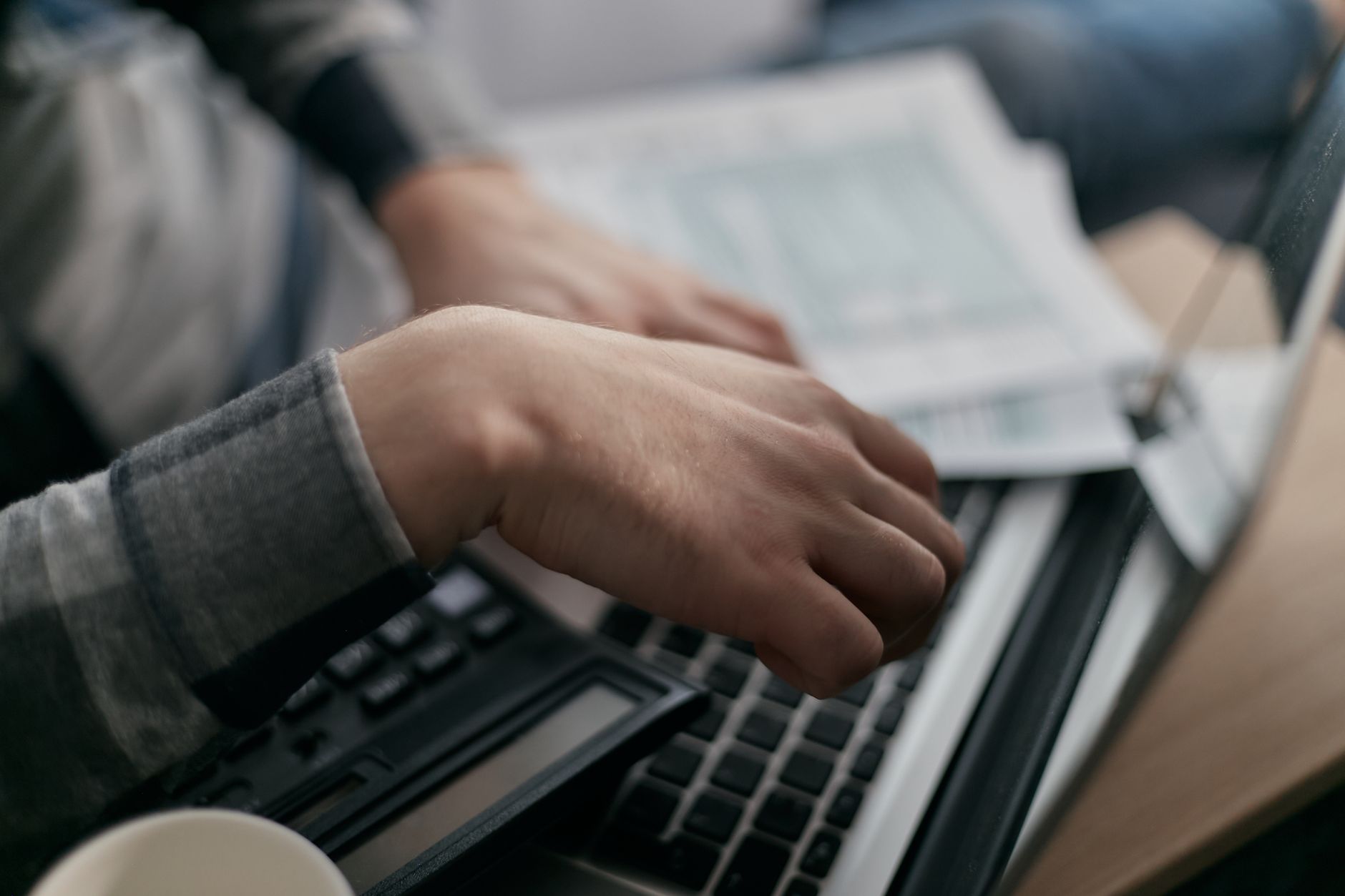 Close-up of a hand on a calculator next to a laptop keyboard, with printed forms on the desk behind
