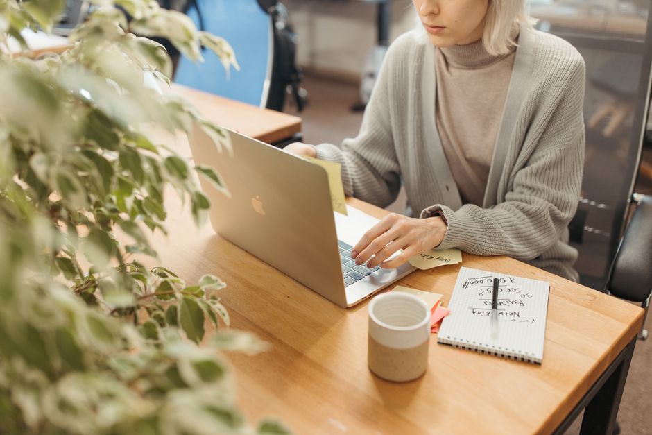 A person typing on a laptop at a wooden desk with a mug and a notepad